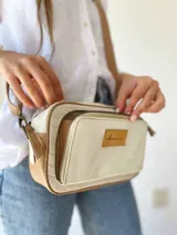 Bandolera rectangular color crudo y marrón, con correa larga regulable, cierre superior con cremallera y bolsillo interior. Fabricada en cuero con forro de tela.