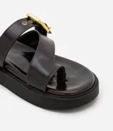 Sandalias planas de PU color marrón oscuro, con tira entre los dedos y tira ancha sobre el empeine con hebilla dorada ajustable.