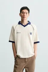 Polo color crema con cuello solapa azul marino, mangas cortas con ribete azul marino, bordado de texto en el pecho y bordado de logo en el abdomen.