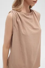 Blusa color beige de poliéster, sin mangas, con cuello asimétrico y detalle de drapeado en los hombros.