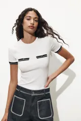 Camiseta blanca de corte recto con cuello redondo y mangas cortas. Presenta dos falsos bolsillos en el delantero con detalle de fornitura a contraste en negro y dorado.