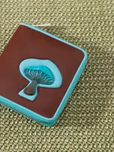 Billetera pequeña de cuero vacuno color bordó con cierre metálico y aplique de hongo. Interior con divisiones para tarjetas y forrería de tejido jacquard.