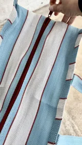 Bolso tipo tote grande con rayas verticales en celeste, blanco y bordó. Tiene doble asa corta en celeste con borde bordó, cierre superior y bolsillo interno.