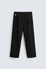 Pantalón negro de corte ancho, con cintura elástica, pliegues frontales, trabilla con hebilla metálica, bolsillos delanteros y traseros de vivo. Cierre frontal con cremallera y botón.
