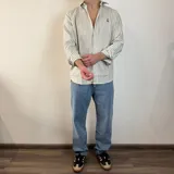 Camisa de manga larga color beige claro, de corte regular, con cuello clásico y cierre frontal con botones. Presenta un pequeño logo bordado en el pecho.
