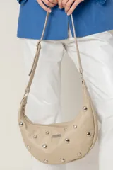 Cartera tipo media luna (hobo bag) de cuero sintético color beige claro, decorada con tachas metálicas plateadas redondas. Posee una correa ajustable para llevar al hombro y cierre superior.