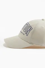 Gorra de sarga color crema con bordado azul marino de la palabra "BOSTON" y debajo "MASSACHUSETTS". Tiene botón en la parte superior, cinta antitranspirante y correa ajustable en la parte trasera.