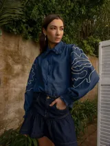 Camisa de denim azul con cuello camisero con volados, cierre frontal con botones, mangas largas abullonadas con puños elastizados y bordado blanco con diseño abstracto en las mangas.