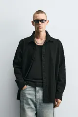 Camisa negra de denim de algodón con textura irregular, corte relaxed fit, cuello solapa, manga larga con puños abotonados y cierre frontal con botones.