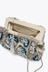 Bolso de mano tipo clutch con cuerpo cubierto de abalorios en tonos beige, azul y gris. Tiene un diseño floral y un cierre imantado. Incluye correa metálica extraíble y bolsillo interior con cremallera.