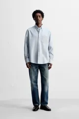Camisa celeste a rayas verticales, de corte regular fit, confeccionada en tejido de algodón. Tiene cuello abotonado, manga larga con puños abotonados, bolsillo de parche en el pecho y cierre frontal con botones.