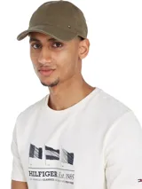 Gorra de béisbol color verde oliva con tejido de espiga, diseño de seis paneles, botón en la parte superior, ojales de ventilación, hebilla de ajuste metálica en la parte trasera y logo esmaltado de Tommy en la parte delantera.