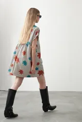 Vestido corto de silueta amplia con corte bajo busto, cuello a la base y mangas cortas. El tejido presenta un estampado de hojas en tonos rojo óxido y turquesa sobre un fondo de rayas verticales blancas y negras.