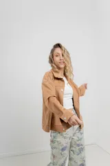 Chaqueta color beige de corte recto, estilo camisa, confeccionada en gamuza con elastano. Presenta cuello camisero, cierre frontal con botones metálicos, bolsillos grandes en el frente y charreteras en los hombros.