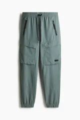 Pantalón jogger deportivo color verde grisáceo, con cintura elástica ajustable con cordón, bolsillos laterales con cierre, bolsillos cargo con solapa y bajos elásticos.