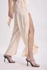 Pantalón de vestir color beige claro, tiro alto, de corte ancho y pierna recta. Presenta un detalle de tela drapeada o solapa larga que cae desde la cintura sobre la pierna izquierda.