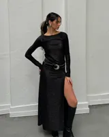 Vestido largo negro de tela fina brillosa, con corte ajustado, mangas largas, cuello redondo y tajo en la pierna. Incluye cinturón de cuero negro con hebilla plateada y tachas.