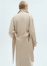Abrigo largo color beige con cuello con solapas, manga larga con puños abotonados, trabillas en los puños, cinturón desmontable con lazada, abertura posterior en el bajo y bolsillos laterales.