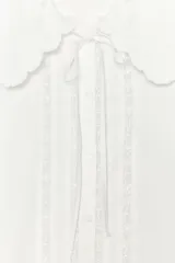 Camisa blanca de cuello bobo con bordados, escote pico con lazada, manga corta abullonada con elástico y cierre frontal con botones.