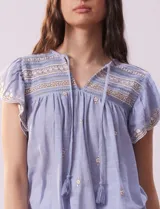 Blusa blanca de algodón con cuello a la base y escote en V con lazo de ajuste. Tiene mangas cortas con terminación amplia y bordados en delantera, mangas y espalda.