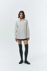 Camisa de manga larga, corte oversized, con cuello clásico y botones frontales. El tejido es de color blanco o muy claro con finas rayas verticales oscuras.