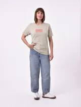 Remera blanca de algodón con cuello redondo y manga corta. Presenta un pequeño bordado en el pecho izquierdo con una figura que parece ser una persona acostada en una tabla de surf o flotador.