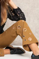 Pantalón de jean color marrón con corte recto, tiro alto y detalle de ojalillos metálicos grandes a lo largo de las piernas.