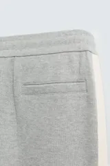 Pantalón de jogging gris con corte relaxed fit, cintura elástica ajustable con cordón interior, bolsillos laterales y traseros de vivo, y banda lateral color crema.