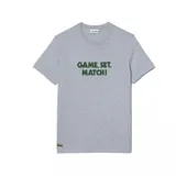 Remera gris de manga corta con cuello redondo y estampado frontal con la frase "GAME, SET, MATCH!" en verde. Logo de Lacoste bordado en el ruedo.