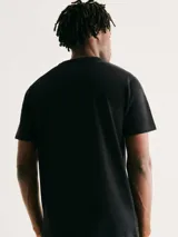 Camiseta blanca de algodón con cuello redondo y mangas cortas. Estampa rectangular con borde negro que contiene la frase "NATURAL ATMOSPHERE" en letras mayúsculas y una imagen de montañas y mar.