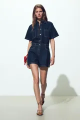 Mono corto de denim azul oscuro, con cuello solapa, manga corta, bolsillos delanteros y traseros, cintura elástica en la espalda y cierre frontal con cremallera y botones metálicos.