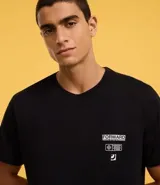 Remera negra de algodón con cuello redondo y mangas cortas. Presenta un estampado con texto y figuras geométricas en el lado superior derecho del frente.