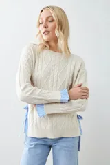 Sweater tejido color crema con diseño de trenzas, corte holgado y camisa celeste a rayas en los laterales.