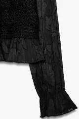 Top negro de cuello redondo y manga larga con puños con volantes. Tiene detalles de crochet en el frente y cierre de botones en la espalda.