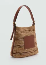 Bolso tipo shopper de paja trenzada color beige con aplique de cuero marrón en el frente. Tiene cierre con botón magnético, bolsillo interior, detalles calados y asa de hombro ajustable con hebillas.