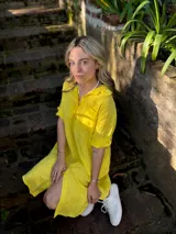 Vestido camisero corto de lino color amarillo, con cuello camisero, mangas cortas con botón, cierre frontal con botones y corte holgado.
