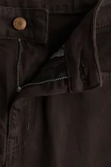 Jean de cinco bolsillos, tiro alto, color marrón oscuro o negro, con piernas anchas y rectas. Presenta cierre frontal con botón y remaches metálicos dorados.