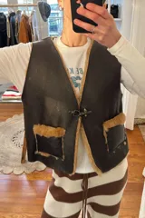 Chaleco marrón de cuero con interior de corderito color crema, cierre con gancho metálico y bolsillos frontales de corderito.