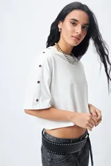 Remera blanca cropped de mangas cortas amplias con ojales metálicos a lo largo de las mangas.