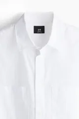 Camisa blanca de popelina de algodón con cuello inglés, tapeta clásica, dos bolsillos superiores abiertos, canesú con pliegue de caja en la espalda, mangas largas con puños abotonados, tapeta de manga con botón y bajo levemente redondeado. Corte holgado.