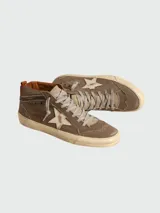 Zapatillas de media caña marca Golden Goose, modelo Mid-Star. Confeccionadas en lona color marrón con efecto desgastado, presentan una estrella de piel blanca en el lateral y detalles de brogue en la puntera y el talón. Cuentan con cordones grises y suela de goma con acabado envejecido.