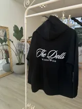 Hoodie negro de corte oversize, con capucha, bolsillo frontal y estampado blanco en la espalda con la inscripción "The Dolls The Dolls VIP CLUB".
