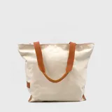 Bolso tipo tote de lona color crudo con detalles en cuero marrón y asas de cuero. Tiene un bolsillo interior con cierre y no está forrado.