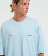 Remera de algodón celeste claro, corte oversized, con cuello redondo y manga corta. Presenta un pequeño texto bordado en azul oscuro en el pecho a cada lado.