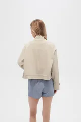 Campera color beige de manga larga, con cuello alto, cierre frontal con cremallera y botones a presión, bolsillos con solapa en el pecho y puños con trabilla ajustable.