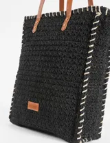 Bolso playero tipo canasto oversize tejido en papiro negro, con costuras decorativas en hilo blanco contrastante y asas de cuero marrón.