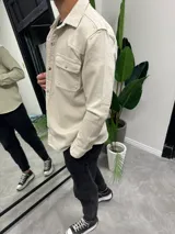 Chaqueta color beige de corte recto, con cuello camisero, cierre frontal con botones metálicos a presión, dos bolsillos con solapa abotonada en el pecho y mangas largas con puños abotonados.