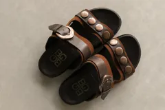Sandalias marrones con doble tira, una con hebilla y otra con apliques metálicos circulares.
