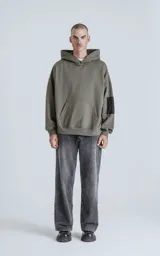 Hoodie color verde militar de rústico 100% algodón, con silueta oversize, capucha forrada y bolsillo tipo bomber en la manga con cierre. Presenta bolsillo canguro frontal.