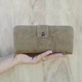 Billetera larga color verde oliva, con cierre de presilla frontal y textura símil cuero desgastado. Presenta un dije metálico redondo con las iniciales "LNS" en azul.